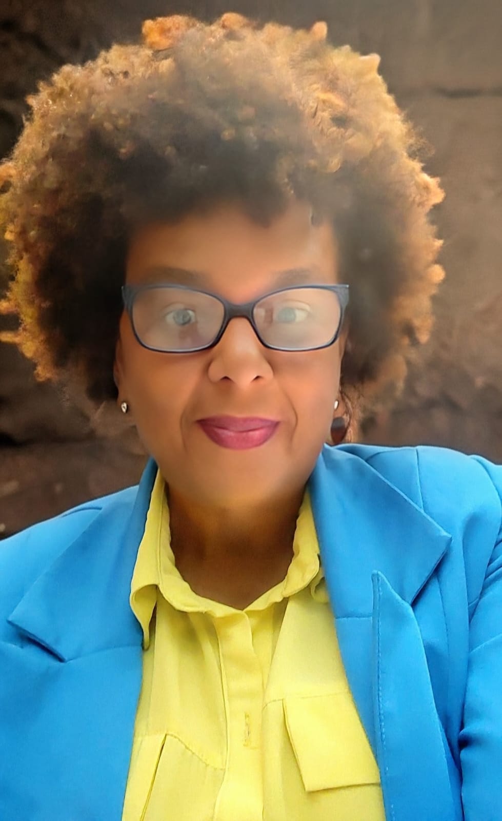 Retrato oficial de Ângela Maria da Silva Gomes, sorrindo, com cabelo black power volumoso e grisalho, óculos escuros de armação azul, brincos pequenos e blusa amarela com blazer azul, sobre fundo de pedra.
