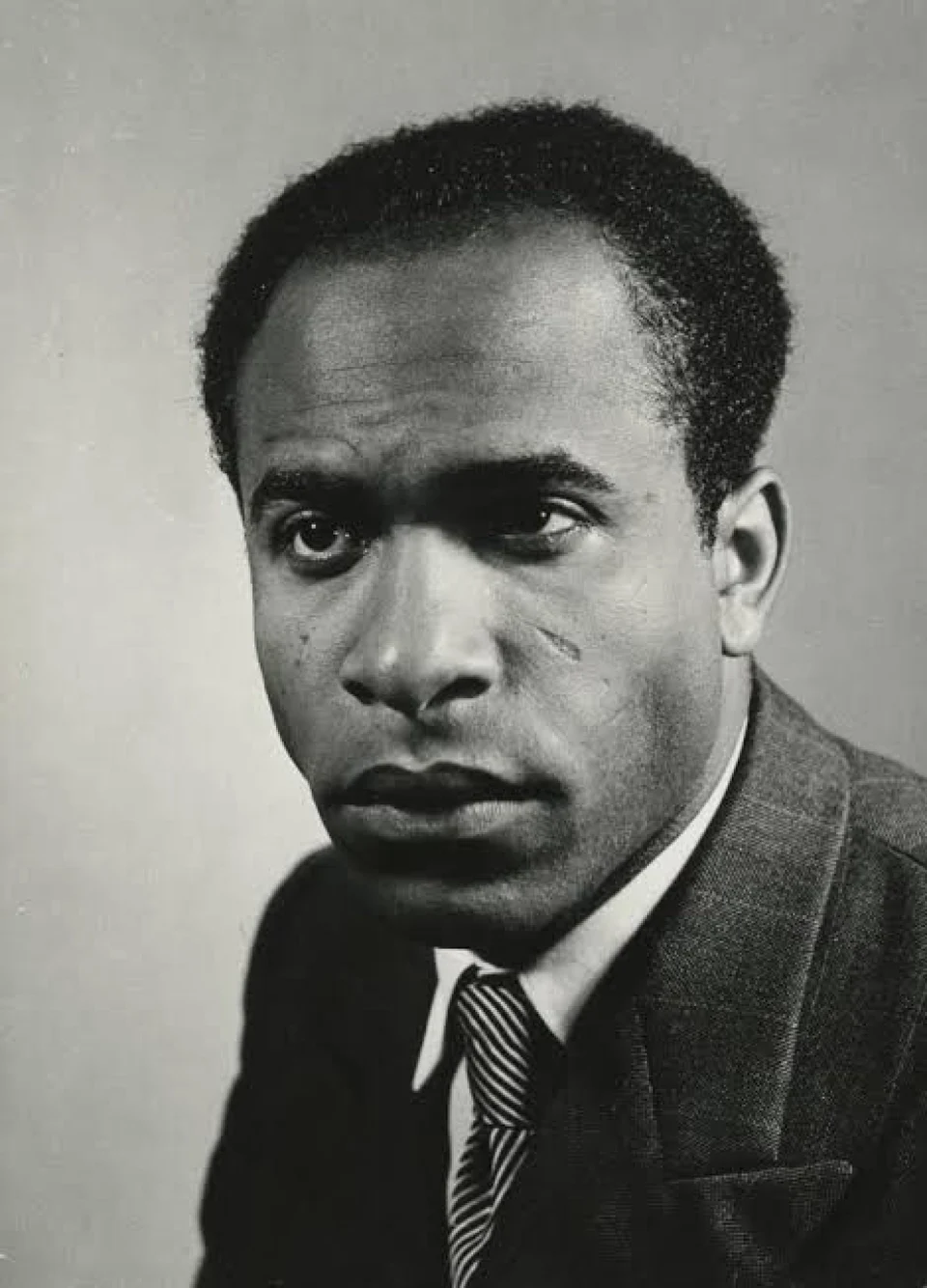 Retrato em preto e branco de Frantz Fanon de terno e gravata listrada, olhando para a câmera.