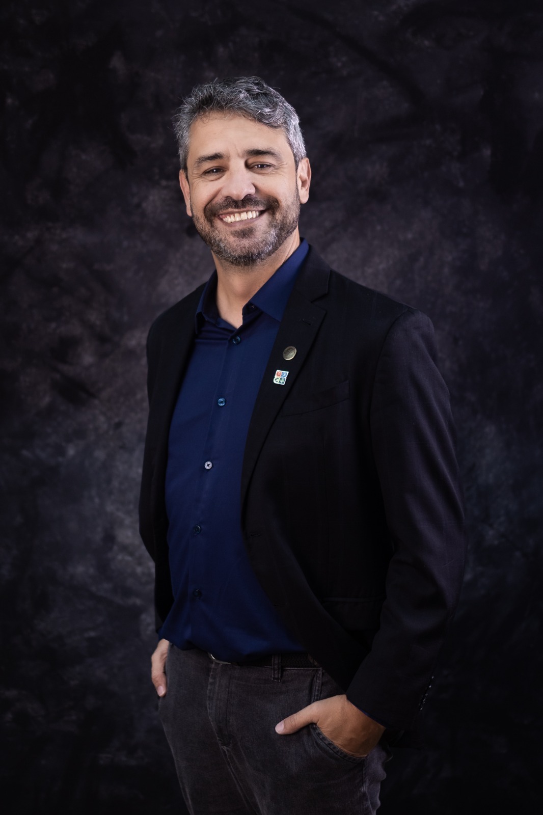 Retrato oficial de Fabiano Gonçalves Guimarães, homem sorridente de cabelo grisalho e barba, usando camisa azul-escura e blazer preto com pins, sobre fundo escuro.