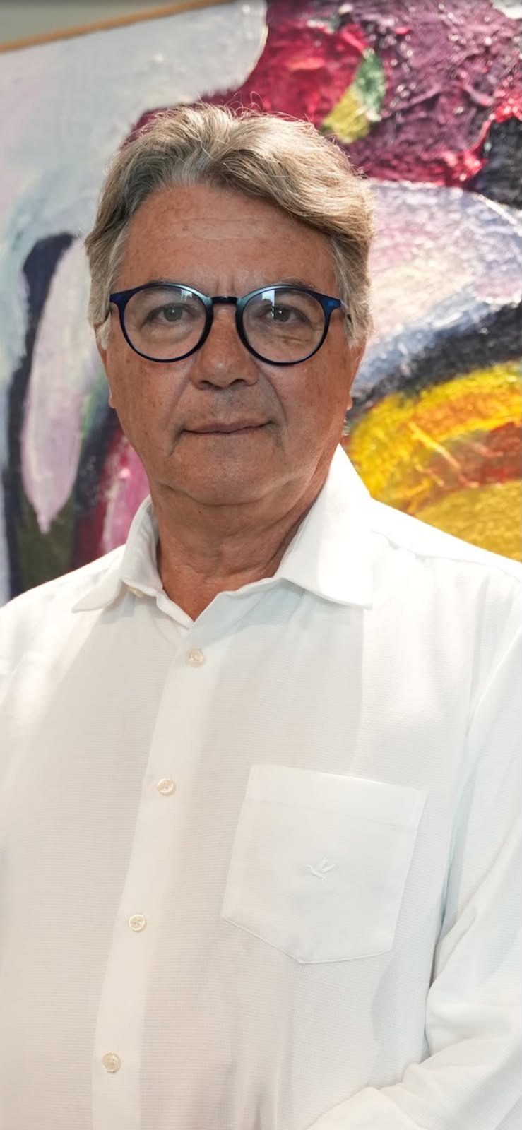 Retrato oficial do debatedor Helvécio Miranda Magalhães Júnior, homem de cabelos grisalhos, óculos de armação arredondada escura e camisa branca, diante de uma pintura abstrata em tons quentes.