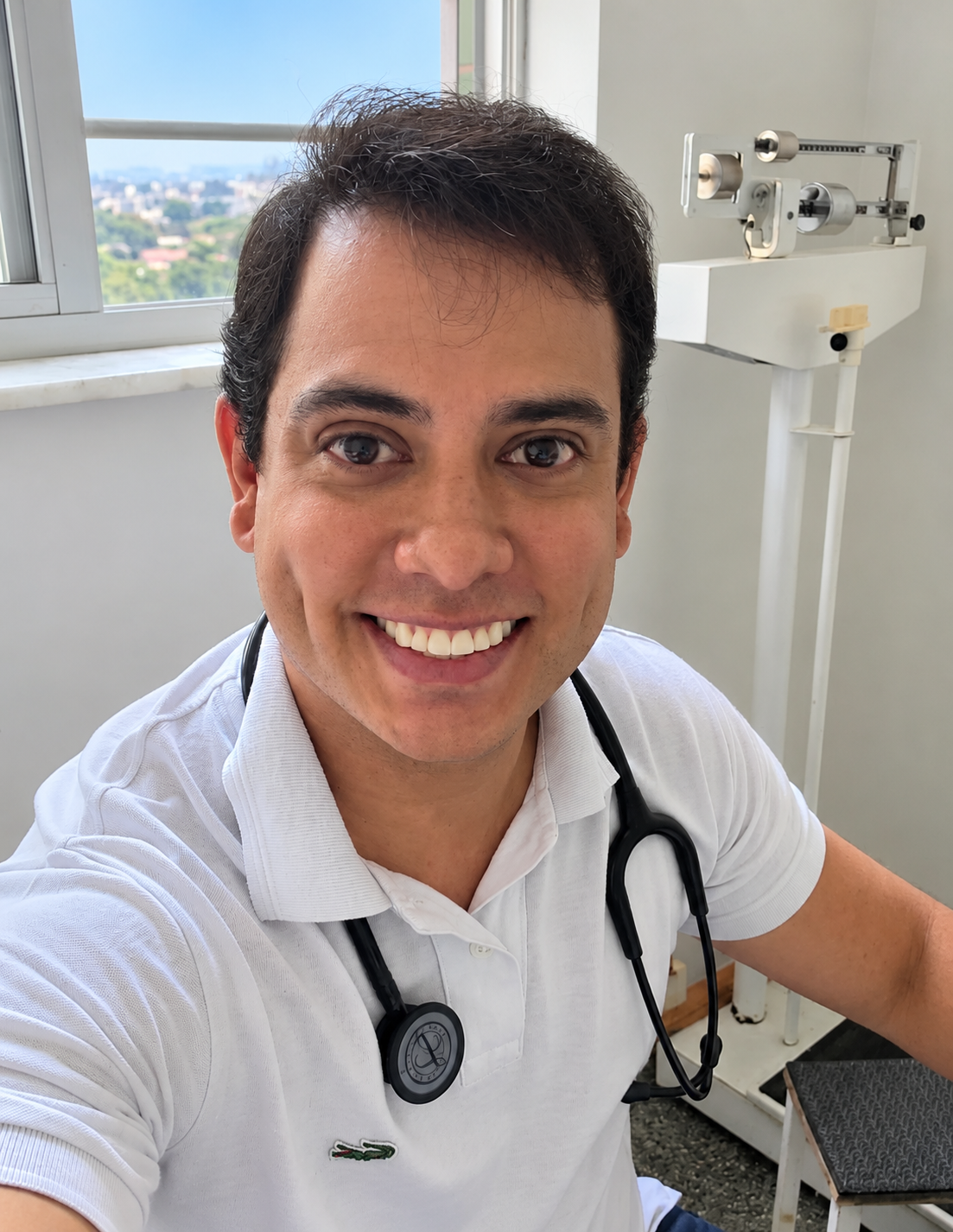 Retrato oficial do debatedor Vitor Corrêa, homem de cabelos escuros ondulados, sorridente, de camisa polo branca com estetoscópio preto no pescoço, em consultório com balança e janela ao fundo.