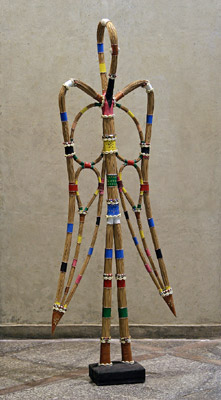 Escultura Iyá Agbá (Mãe Ancestral), de Mestre Didi: formas hibridas com pernas alongadas, estruturas semelhantes a asas e formas em gota, em fibras trançadas com faixas de contas coloridas, sobre base escura.