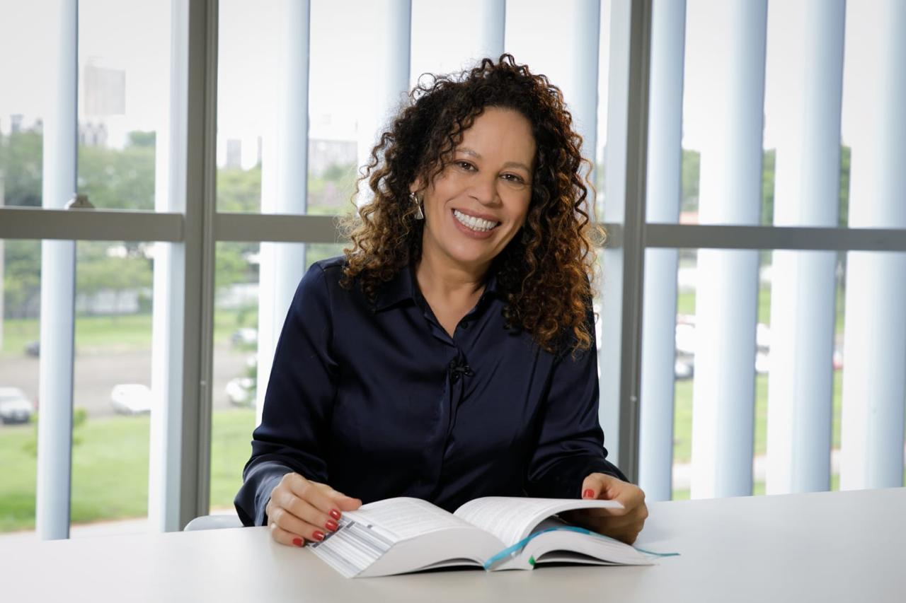 Retrato oficial de Edilene Lobo, sorrindo, sentada a uma mesa com um livro aberto, cabelo cacheado e camisa azul-escura, diante de uma janela.