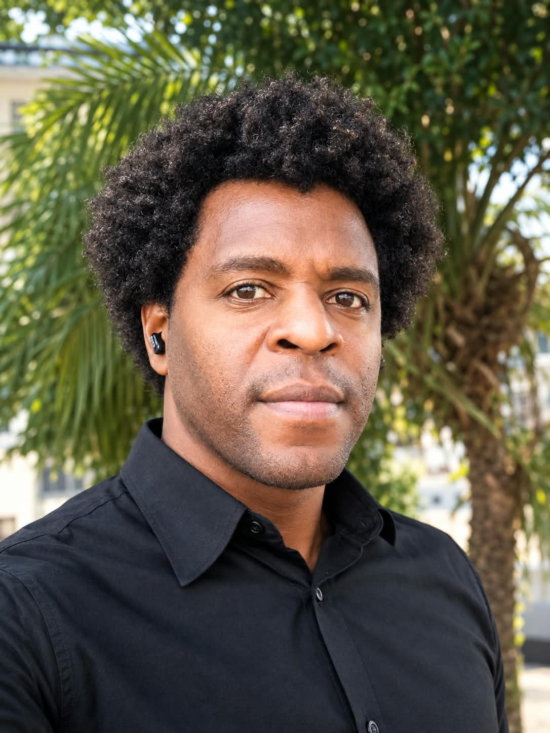 Retrato fotográfico de Robson Julio Romualdo, homem negro de cabelo black power volumoso, camisa preta, diante de árvores.
