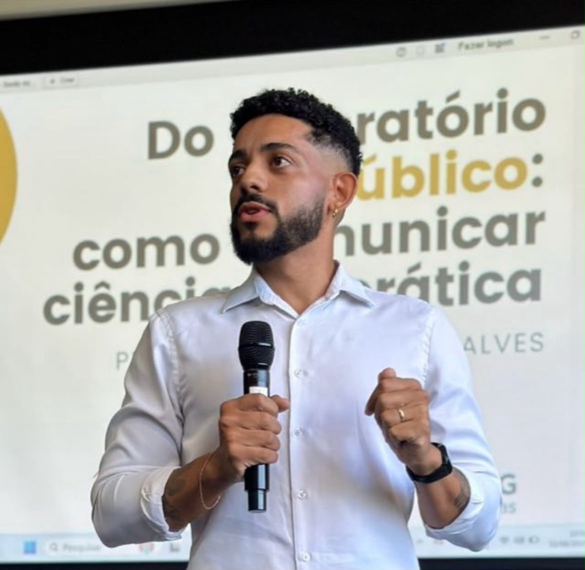 Retrato oficial de Lucas Gonçalves, homem de cabelo curto e barba cheia, falando ao microfone durante uma palestra, com camisa branca e fundo com projeção de slides.
