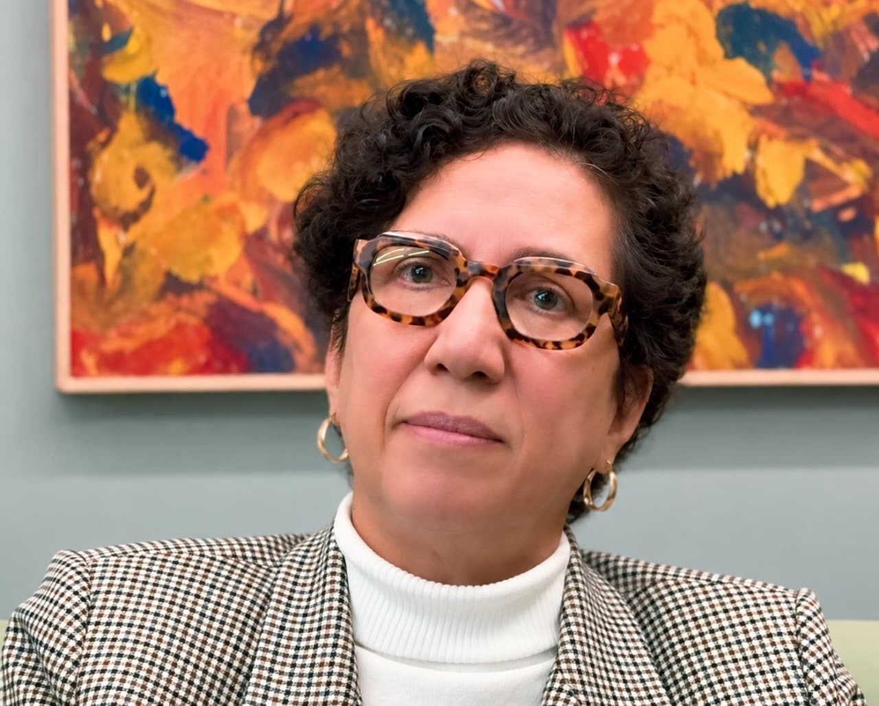Retrato oficial da debatedora Maria do Carmo, mulher de cabelos curtos cacheados escuros, óculos de armação quadriculada, brincos argola dourados, blazer quadriculado e blusa branca de gola alta, diante de uma pintura abstrata em tons quentes.