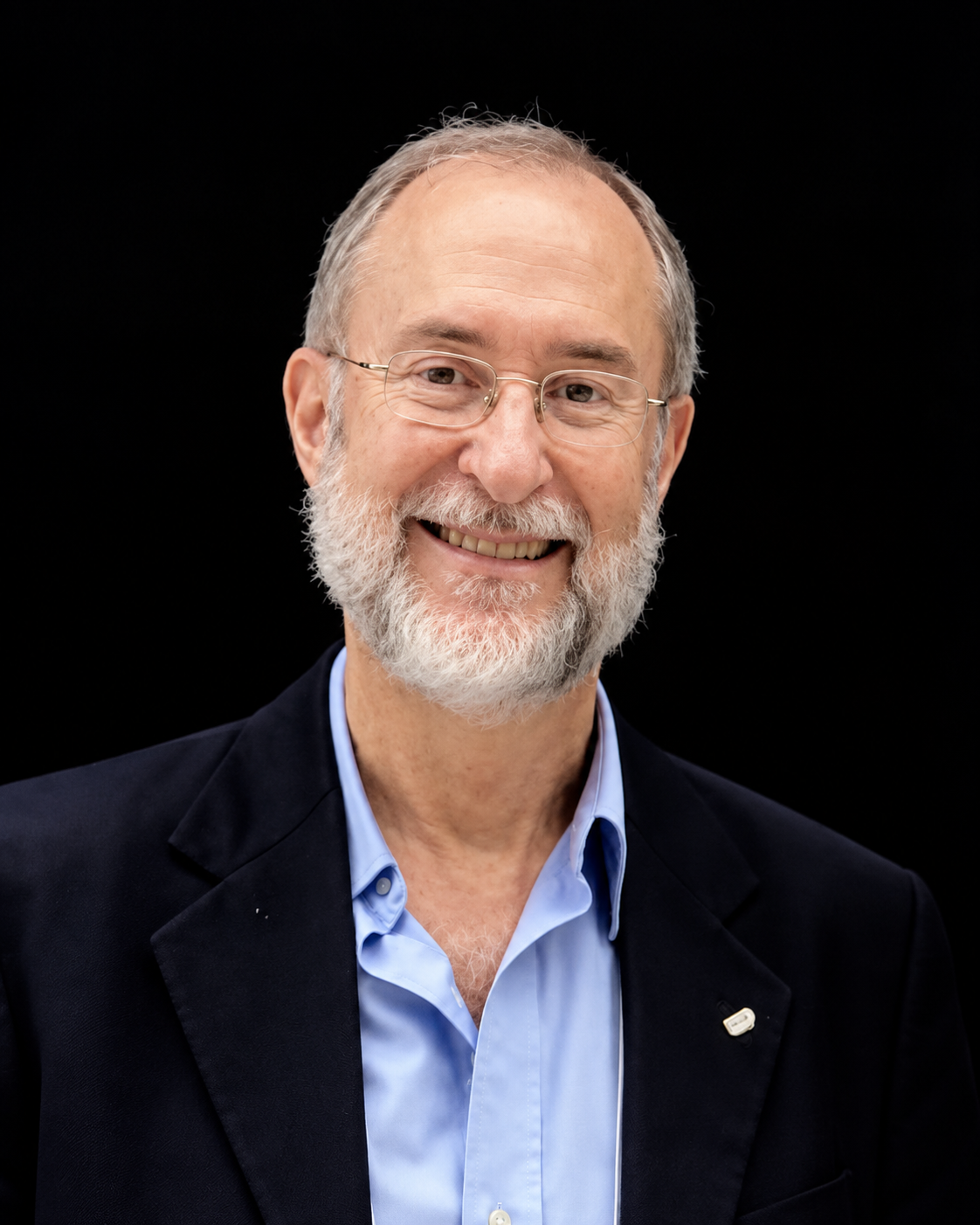 Retrato de António Mauro Saraiva, sorrindo, com cabelos grisalhos, barba branca, óculos de armação fina, camisa azul-clara e blazer escuro, sobre fundo escuro.