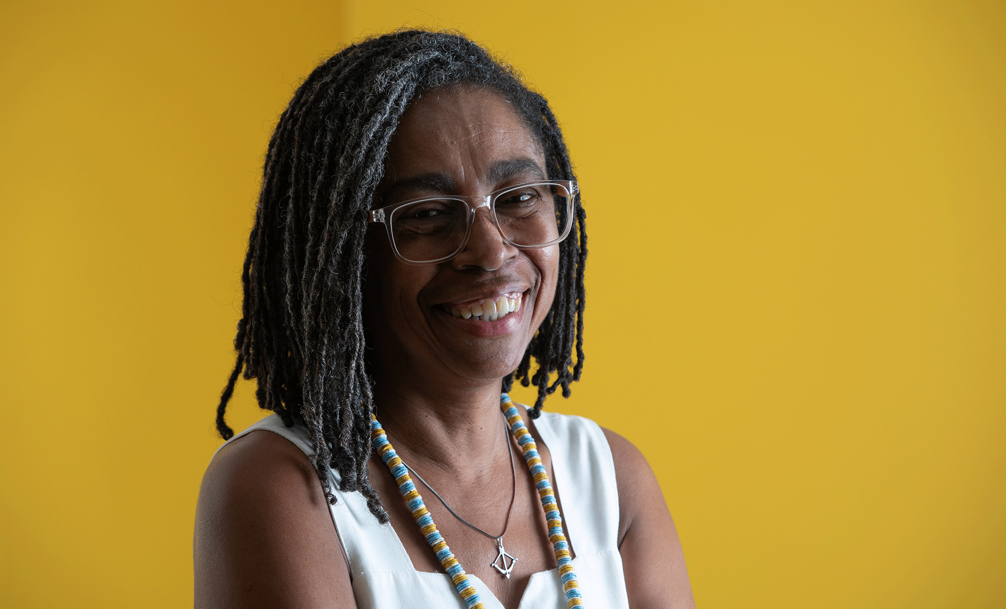 Fotografia de Jurema Werneck sorrindo diante de fundo amarelo, com óculos, tranças e colar de contas.