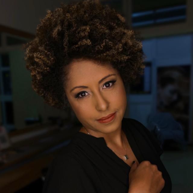 Retrato oficial de Renata Mendonça, com cabelo black power volumoso, brincos dourados, colar e blusa preta, sobre fundo escuro.