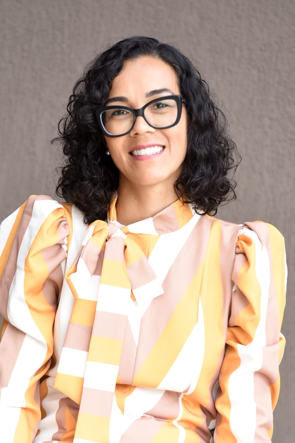 Retrato oficial da debatedora Marina Abreu Corradi Cruz, mulher de cabelos escuros ondulados na altura dos ombros, óculos de armação escura, sorrindo, de blusa listrada em tons de amarelo, bege e branco, sobre fundo cinza.