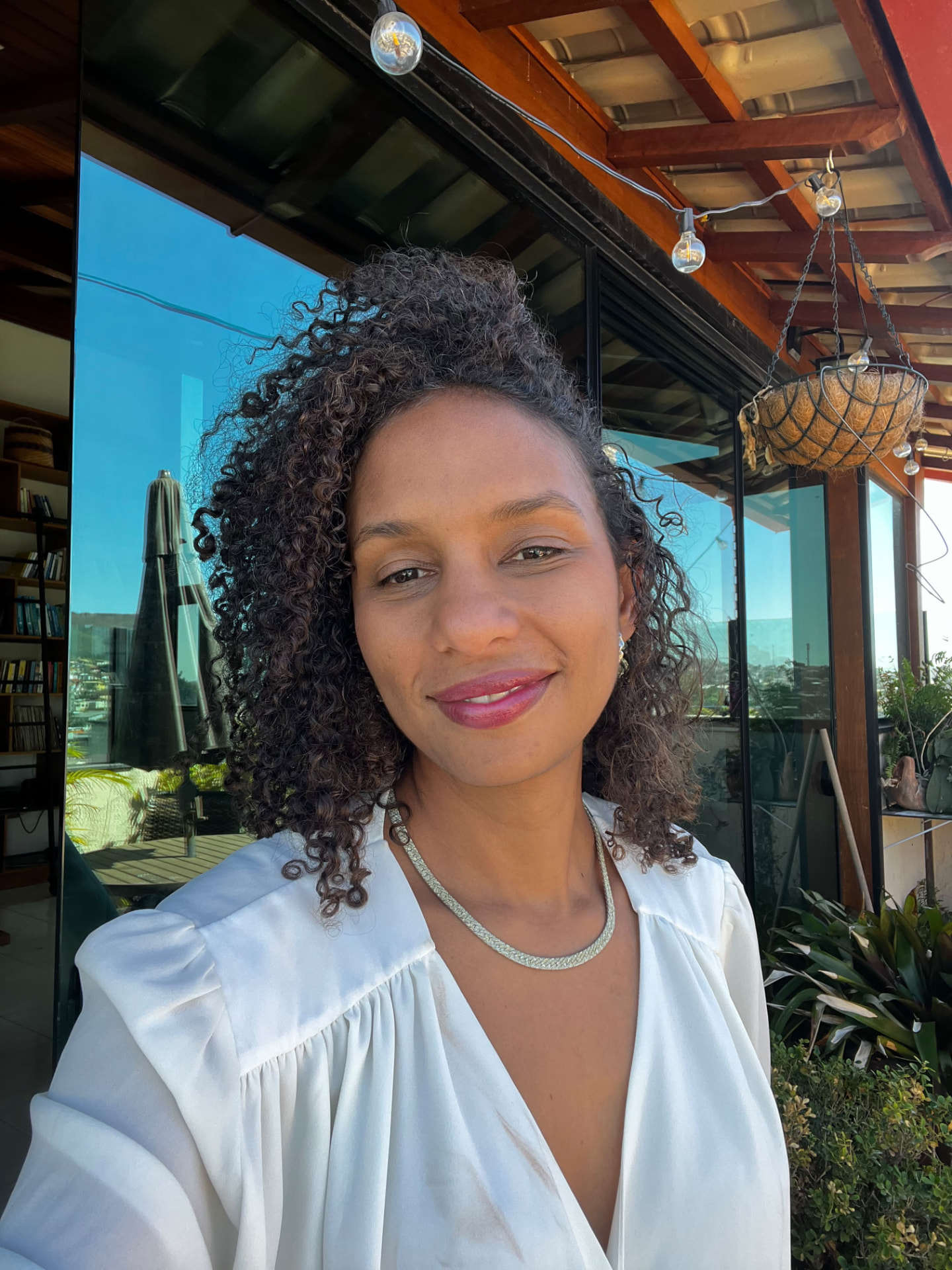 Retrato oficial da debatedora Clarissa Santos Lages, mulher de cabelos escuros cacheados na altura dos ombros, sorrindo, de blusa branca com colar prateado, em varanda com plantas e vidros ao fundo.