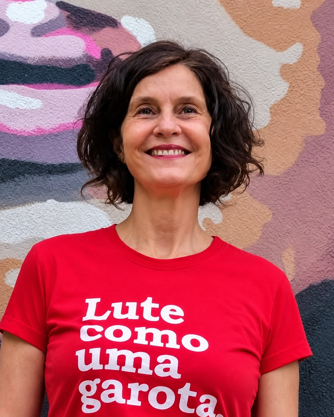 Retrato de Sônia Lansky, sorrindo, com cabelo curto escuro, camiseta vermelha com a inscrição "Lute como uma garota", diante de um mural colorido.