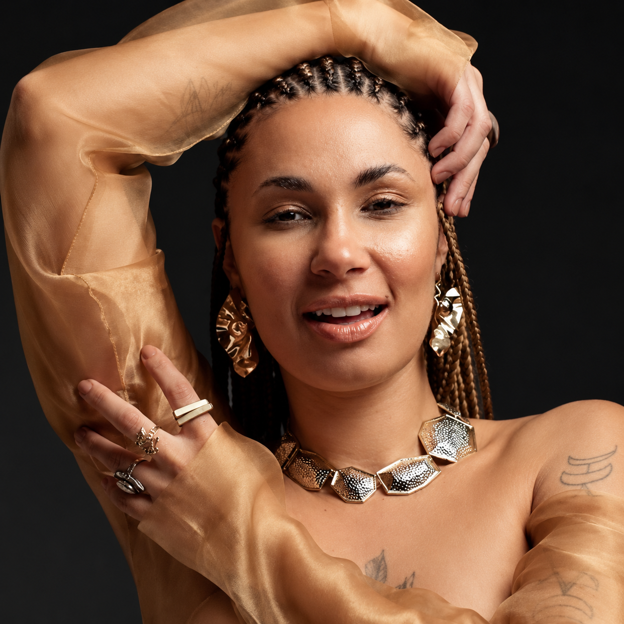 Retrato fotográfico de Maíra Baldaia, de tranças, brincos, colar e anéis dourados e vestido transparente cor de ouro, sorrindo diante de um fundo escuro.