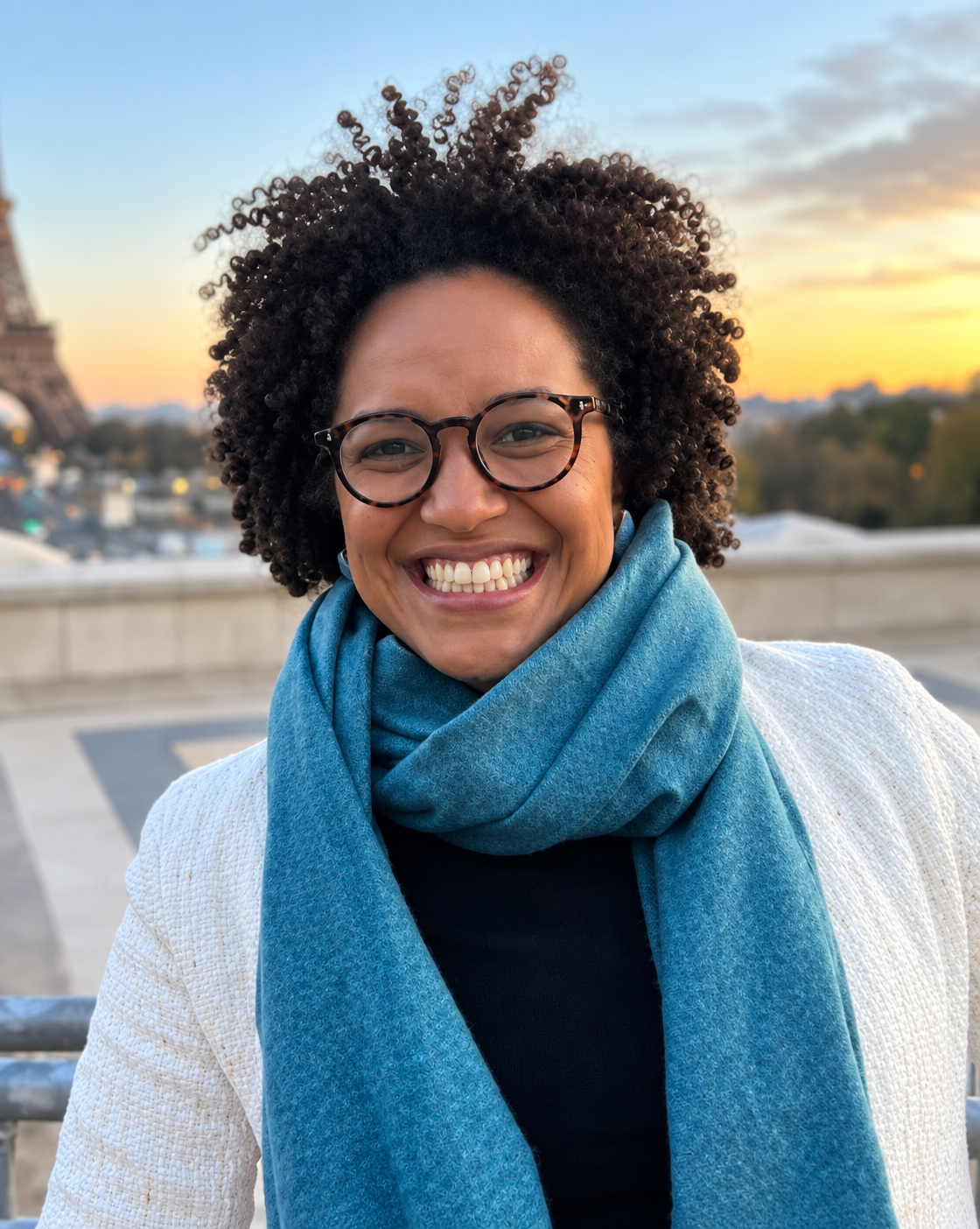 Retrato oficial de Lorena Luiza Chagas Lemos, sorrindo, com cabelo black power, óculos, cachecol azul e casaco claro, em ambiente externo ao entardecer.