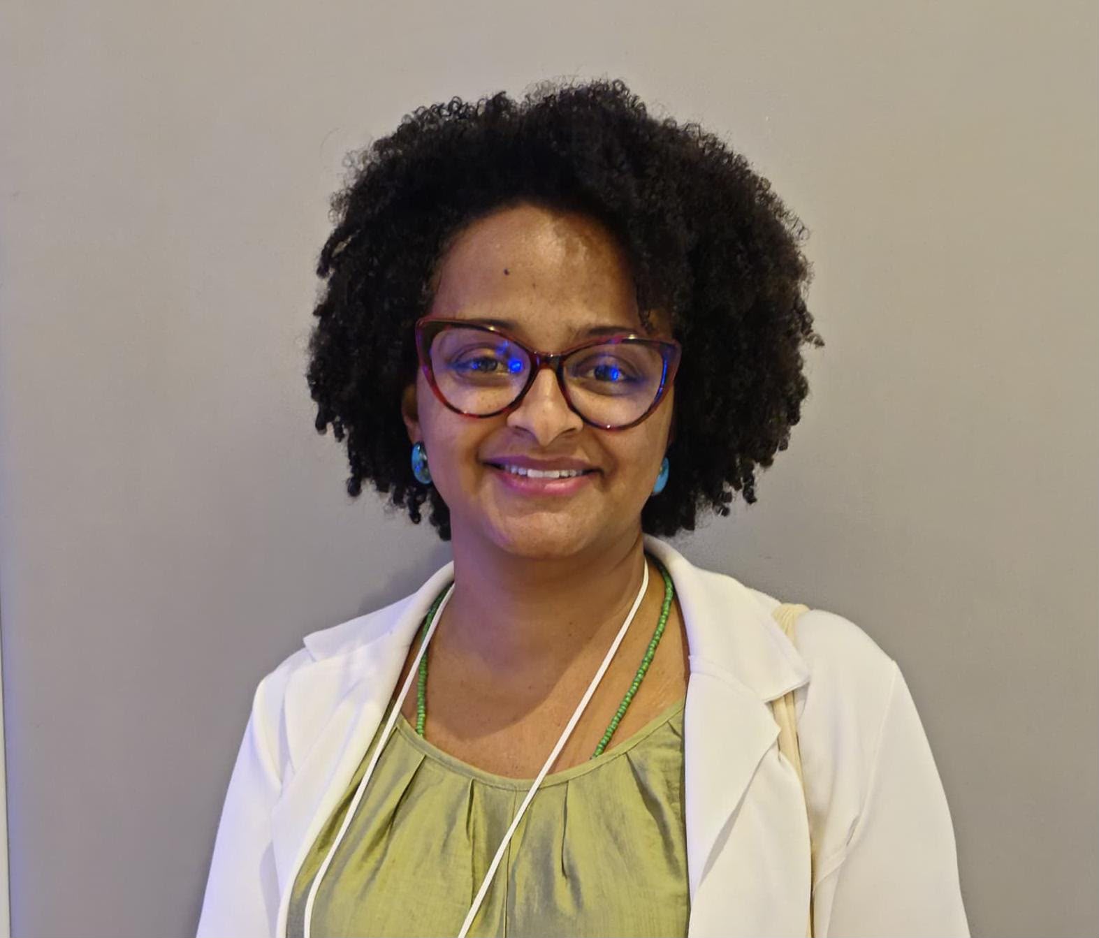 Retrato oficial de Ana Cristina de Oliveira Costa, sorrindo, com cabelo cacheado volumoso, óculos de armação vinho, brincos azuis, blusa verde-oliva e blazer branco, sobre fundo neutro.