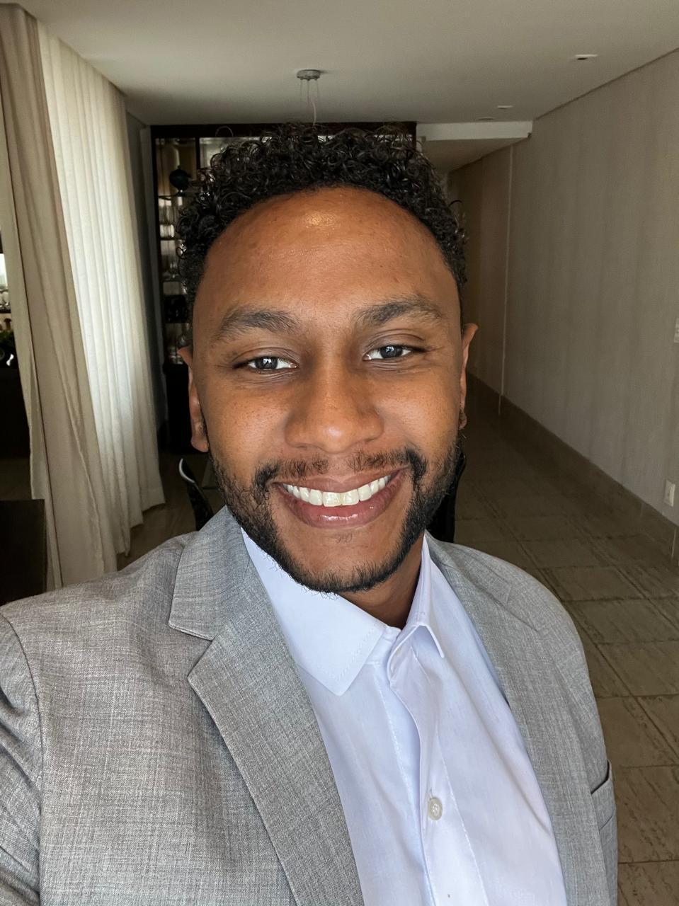 Retrato oficial de Gustavo Mariano Rodrigues Santos, sorrindo, com cabelo cacheado curto, barba cerrada, camisa social azul-clara e blazer cinza-claro, em ambiente interno.
