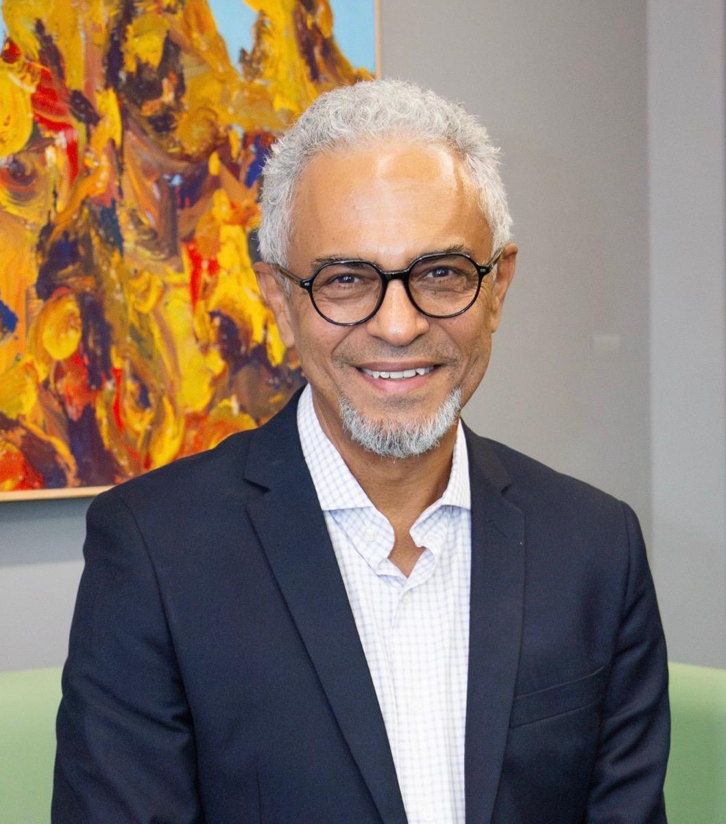Retrato oficial do debatedor, homem de cabelos grisalhos, óculos de armação arredondada, blazer azul-marinho e camisa quadriculada clara, sorrindo diante de uma pintura abstrata em tons quentes.